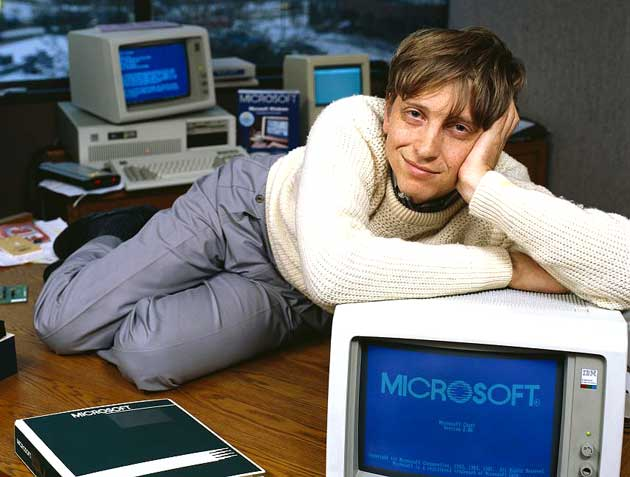 A young Bill Gates reclining across a desk beside 1980s PCs running Microsoft software